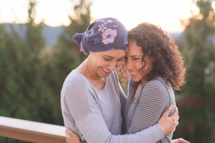 A beautiful young ethnic woman fighting cancer and wearing a head wrap embraces her sister. They are tightly holding each other and she is looking down and smiling. Her sister is also smiling. They are standing outdoors and there are mountains and trees in the background.